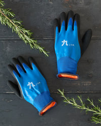 Pair of blue gardening gloves with black cuffs on a dark wooden surface with greenery.