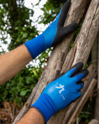 Person wearing blue gardening gloves with black accents on a wooden branch outdoors.