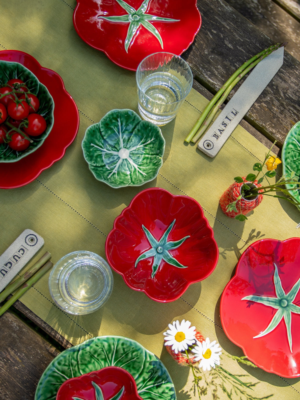 Colorful ceramic plates with tomato design on a wooden table with glasses and flowers.