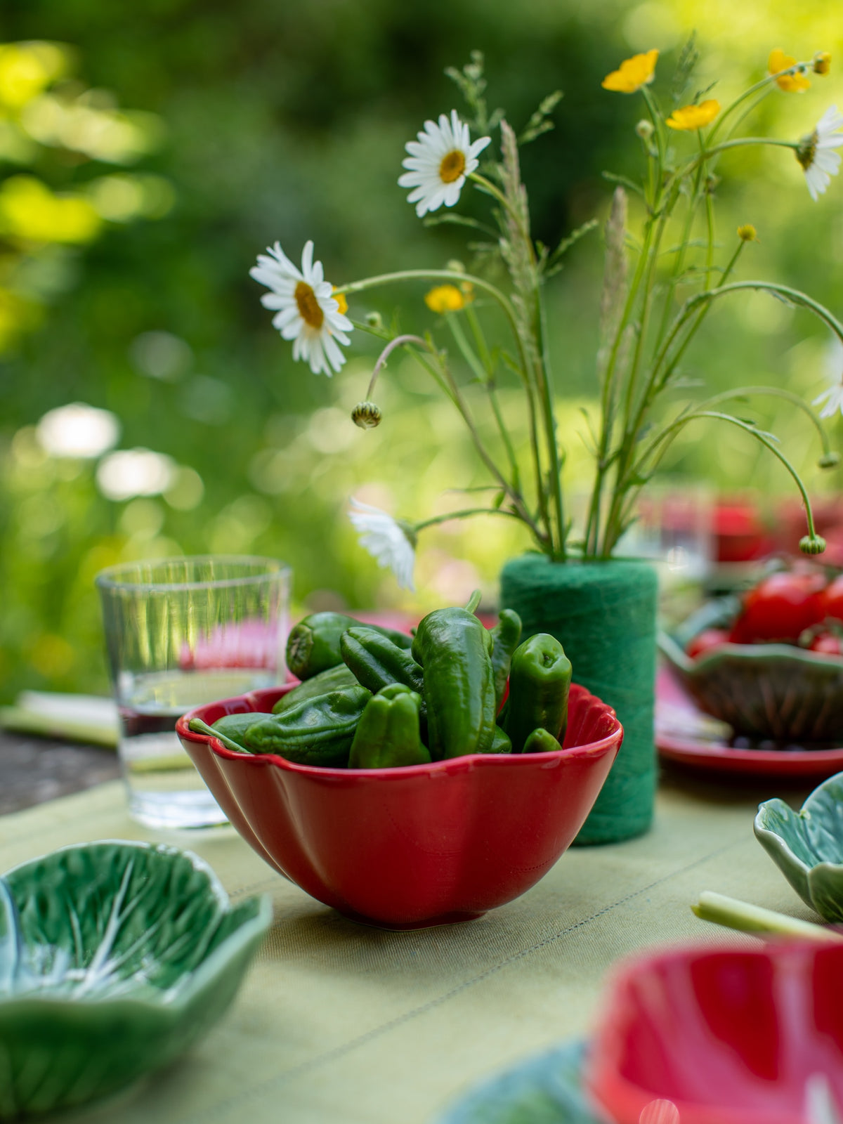 Outdoor setting with a table featuring a red bowl of green peppers, a glass of water, and a vase with flowers.
