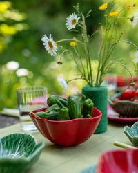Outdoor setting with a table featuring a red bowl of green peppers, a glass of water, and a vase with flowers.