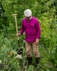 Man in a garden wearing a magenta shirt and a flat cap, holding a gardening tool.