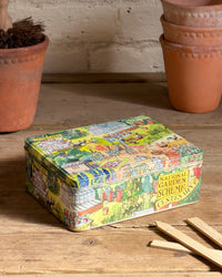 A square metal tin celebrating the centenary of the National Gardens Scheme with a colourful vintage-style garden illustration on the lid, placed on a wooden table with terracotta pots in the background.