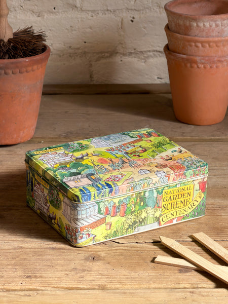 A square metal tin celebrating the centenary of the National Gardens Scheme with a colourful vintage-style garden illustration on the lid, placed on a wooden table with terracotta pots in the background.