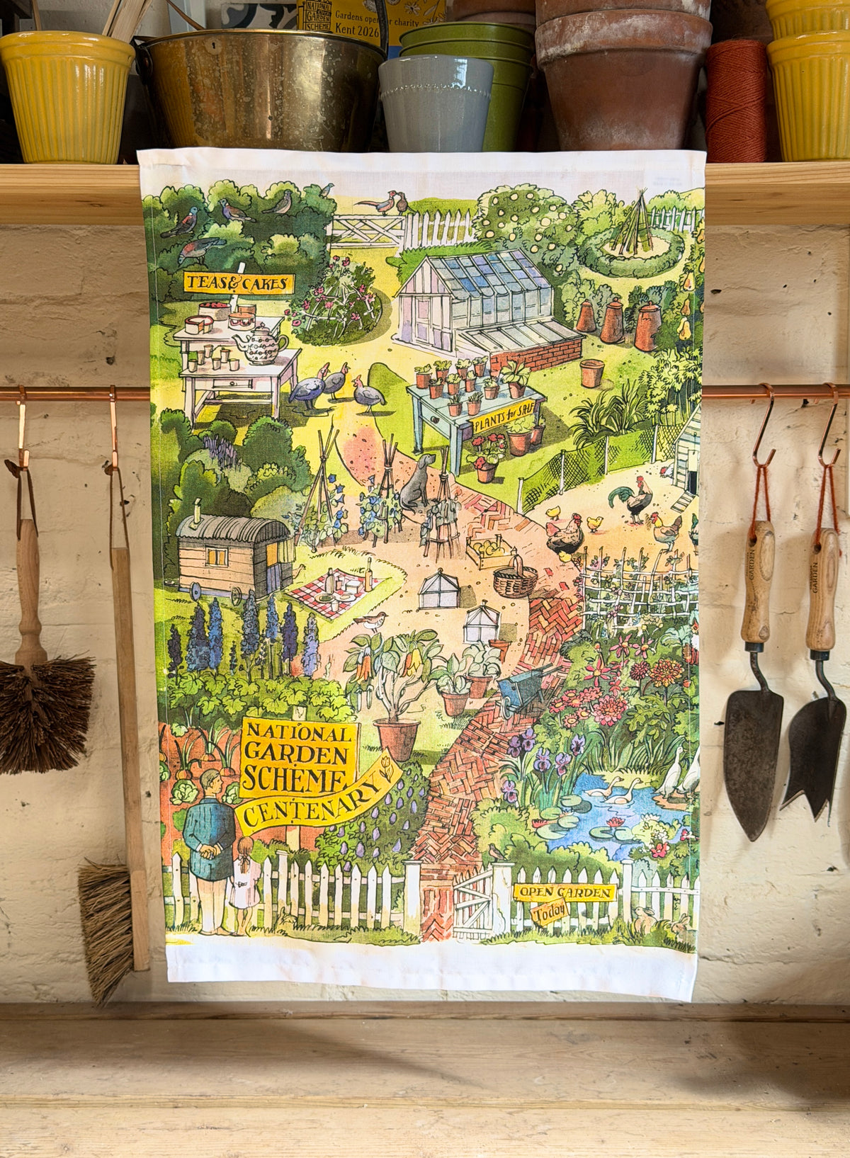 A rectangular tea towel with a colorful illustrated garden scene hangs on a wooden shelf. The towel features a whimsical landscape with a greenhouse, flowers, chickens, a pond, and garden tools.