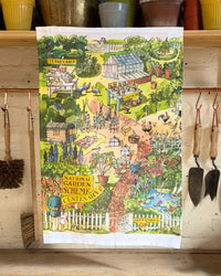 A rectangular tea towel with a colorful illustrated garden scene hangs on a wooden shelf. The towel features a whimsical landscape with a greenhouse, flowers, chickens, a pond, and garden tools.