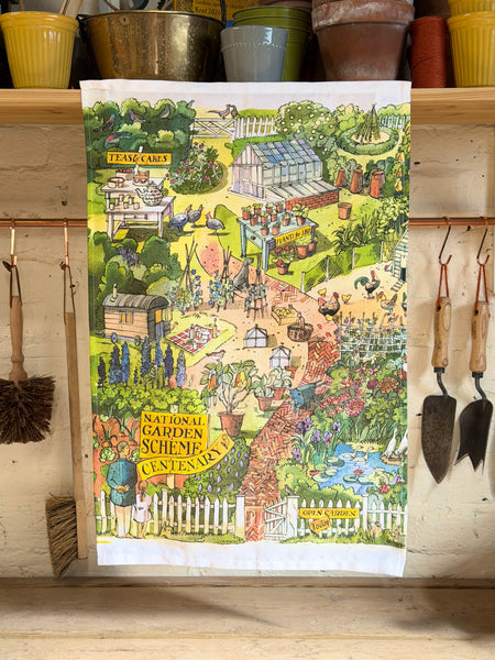A rectangular tea towel with a colorful illustrated garden scene hangs on a wooden shelf. The towel features a whimsical landscape with a greenhouse, flowers, chickens, a pond, and garden tools.