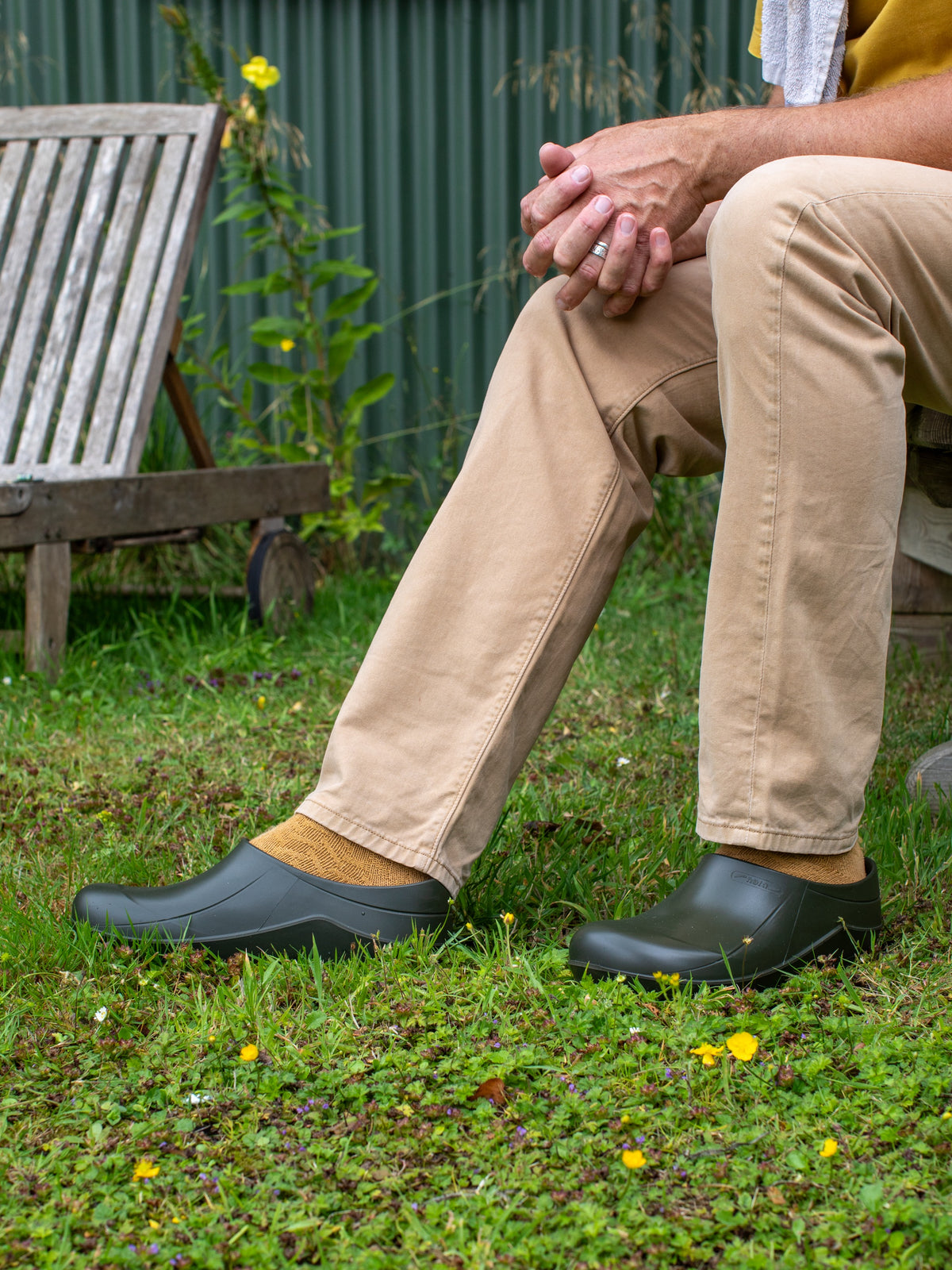 Person wearing green clogs sitting on grass with a garden background