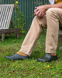 Person wearing green clogs sitting on grass with a garden background