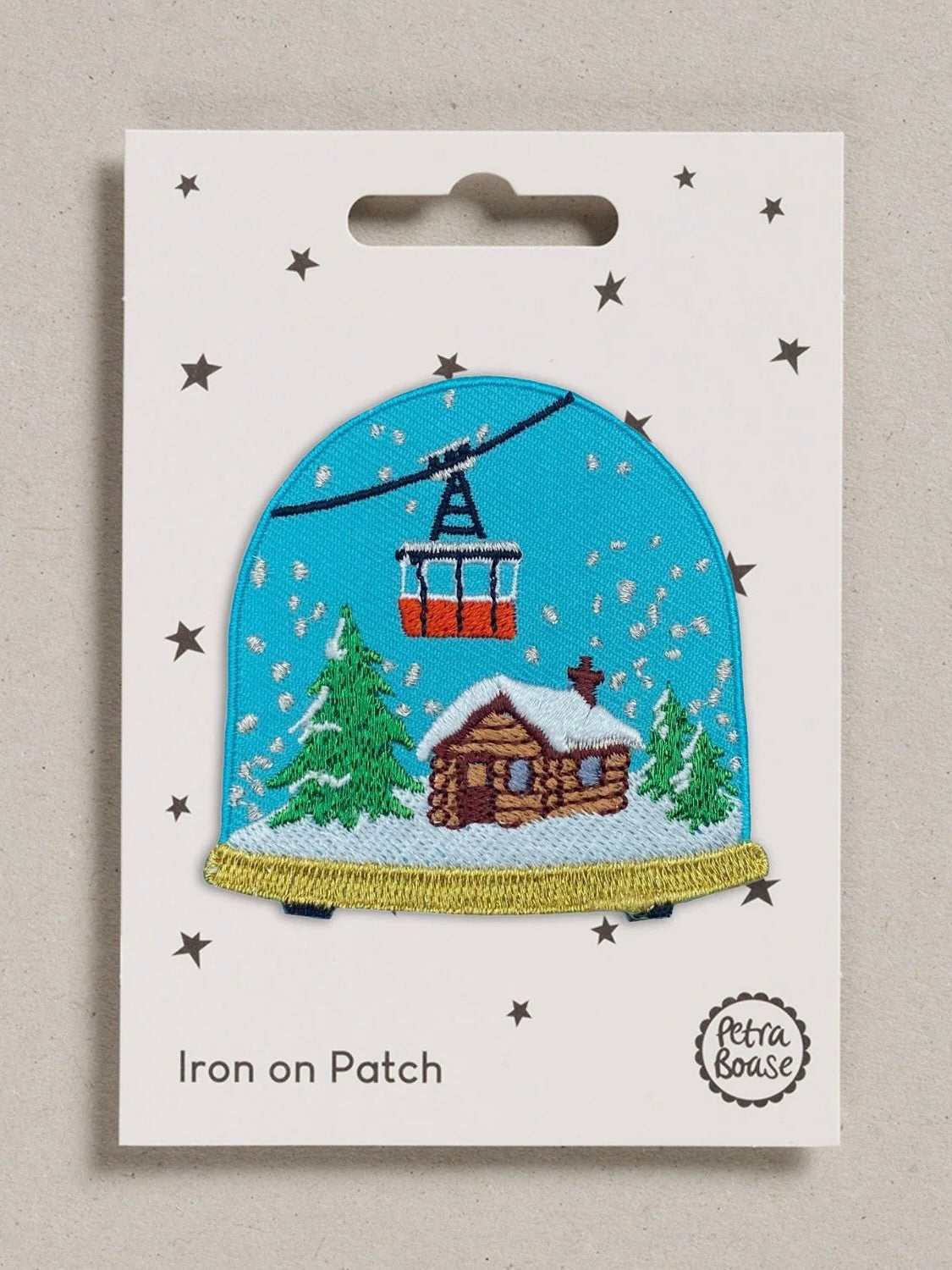 An embroidered iron-on patch depicting a snowy mountain cabin under a cable car, mounted on a white backing card with star details and the brand name Petra Boase.