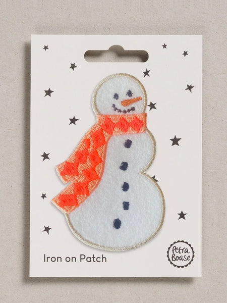 A snowman iron-on patch on a white backing card with star details, photographed against a neutral textured gray background. The patch features a white body, an orange patterned scarf, and dark blue buttons.