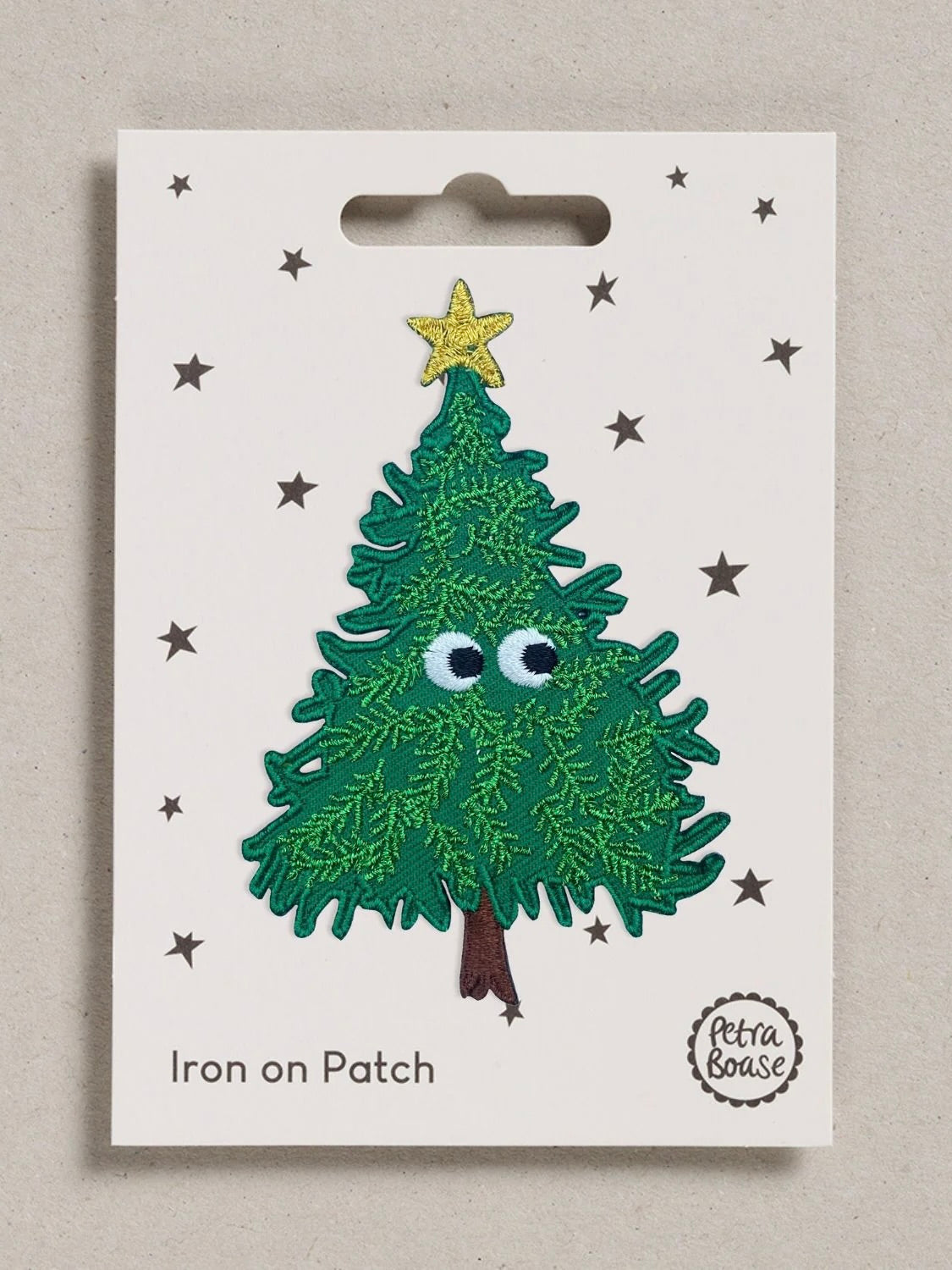 A Christmas tree iron-on patch displayed on its original white backing card. The patch features a green textured tree with googly eyes, a yellow star on top, and a brown trunk.