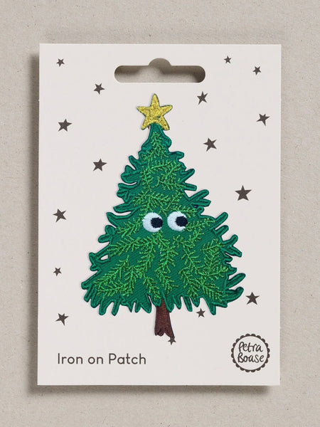 A Christmas tree iron-on patch displayed on its original white backing card. The patch features a green textured tree with googly eyes, a yellow star on top, and a brown trunk.